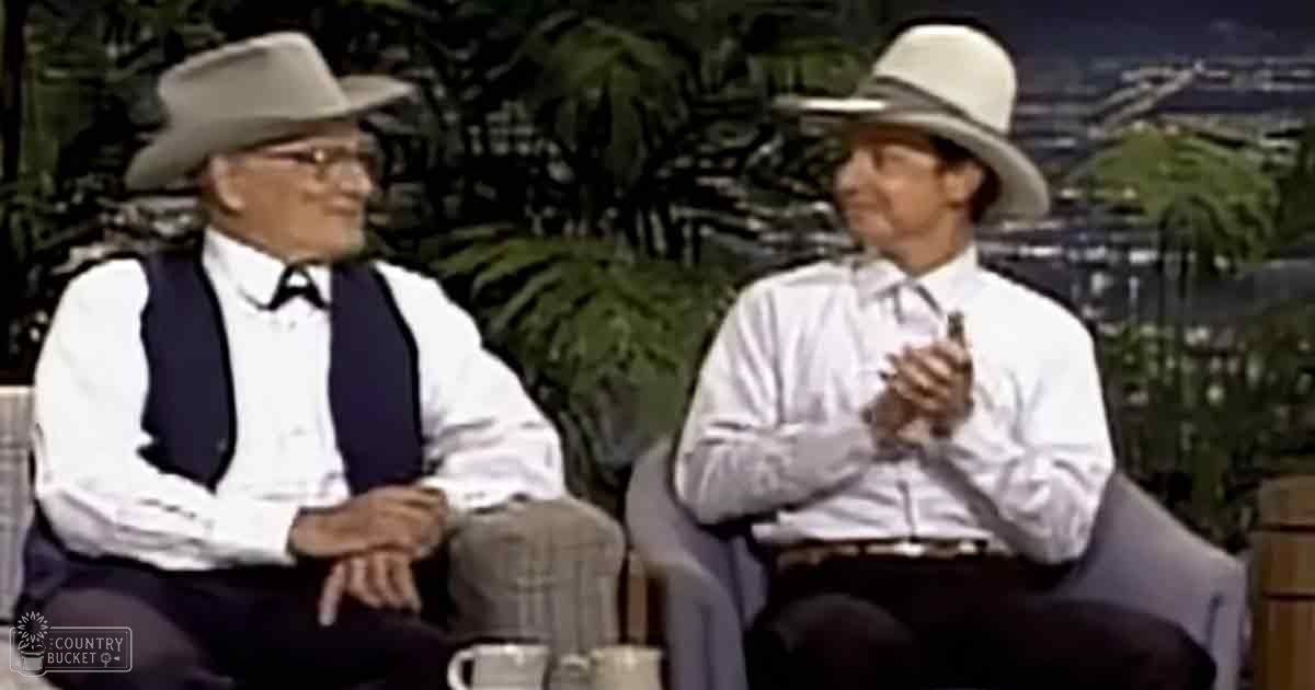 Two older adult males wearing matching outfits with white shirts, dark pants, and beige cowboy hats in a studio setting with light gray upholstered chairs and plants.