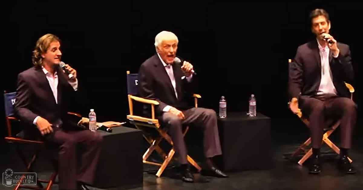 Panel discussion with three adult males in a formal indoor setting, each wearing suits and holding microphones.