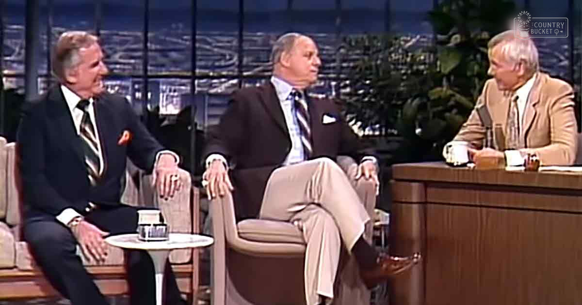 Three older adult males in formal attire seated on a talk show set with an urban backdrop and warm lighting.