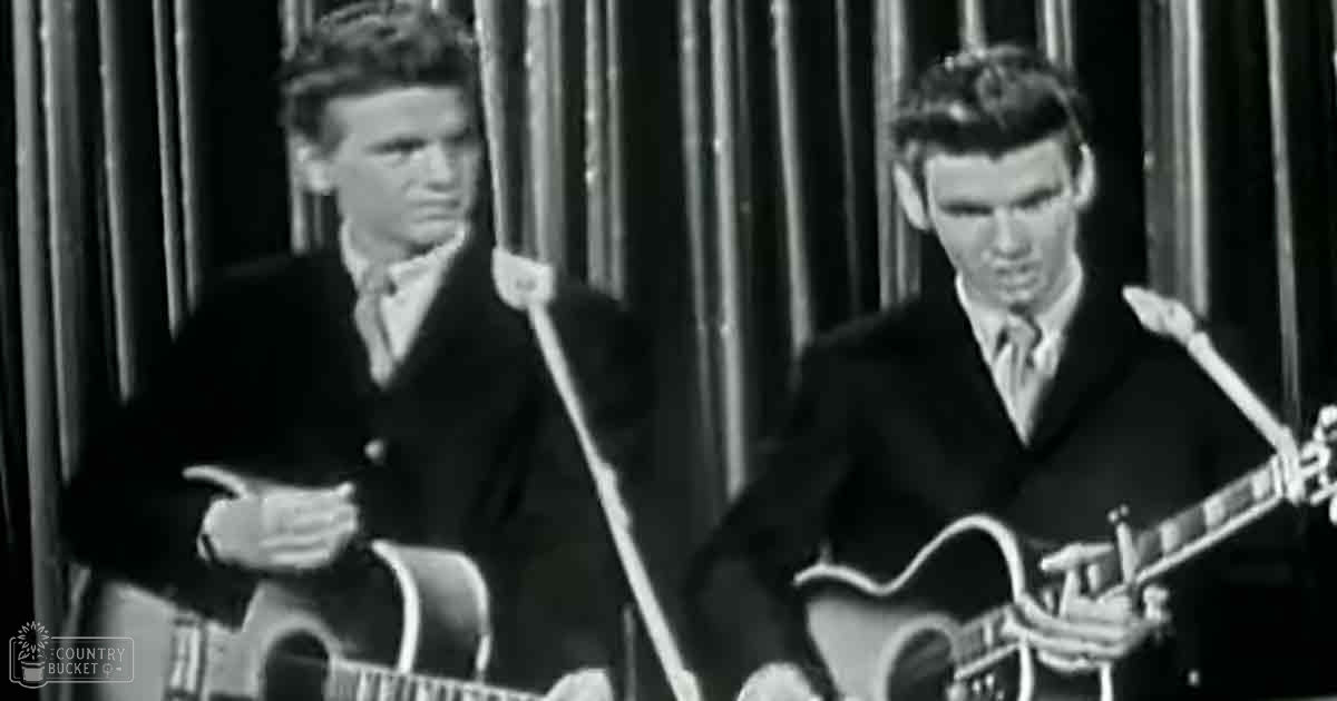 Two adult males wearing dark suits with notched lapels, performing music in a mid-20th century studio setting with microphones and acoustic guitars.