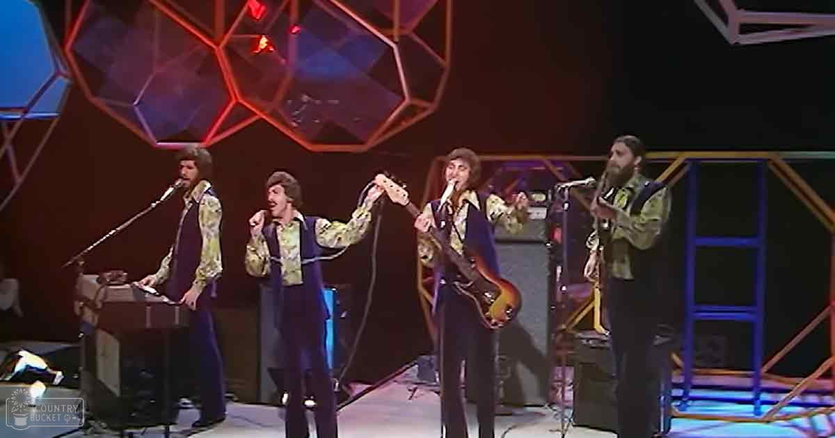 A musical performance from the 1970s with four adult male singers on stage, playing electric guitars and keyboards under vibrant red and purple light patterns projected onto a screen behind them.