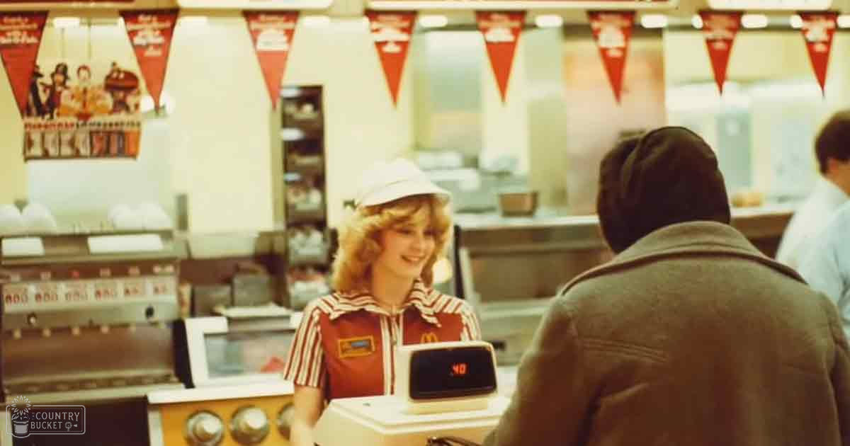 A young adult female cashier at McDonald’s wearing a red Adidas tracksuit, interacting with customers inside an indoor fast-food restaurant setting.