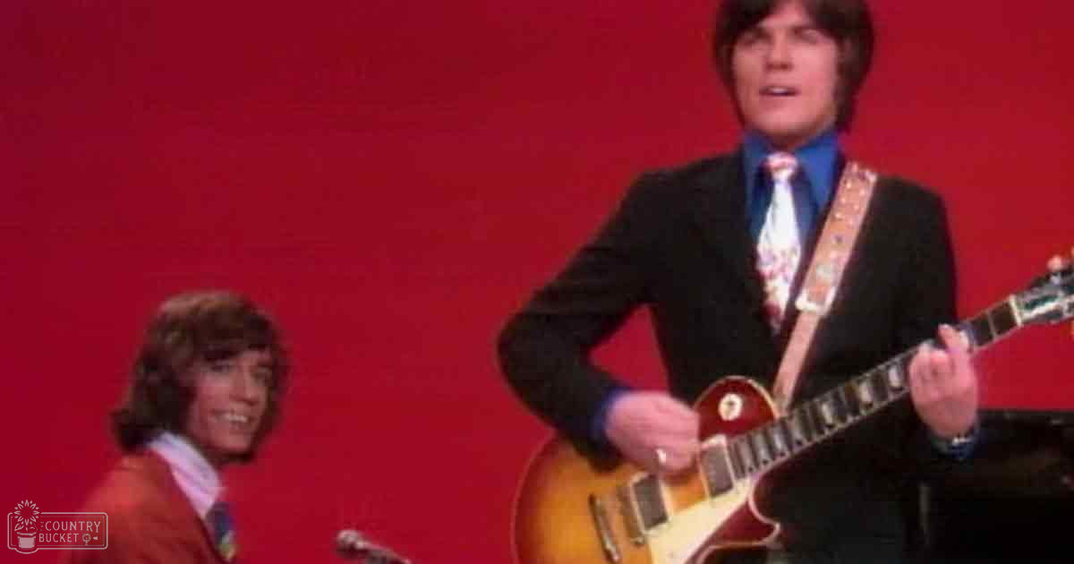 A young adult male, dressed in a dark suit with a patterned tie, plays an electric red guitar against a plain red background. A child or teenager wearing a red blazer stands behind him.