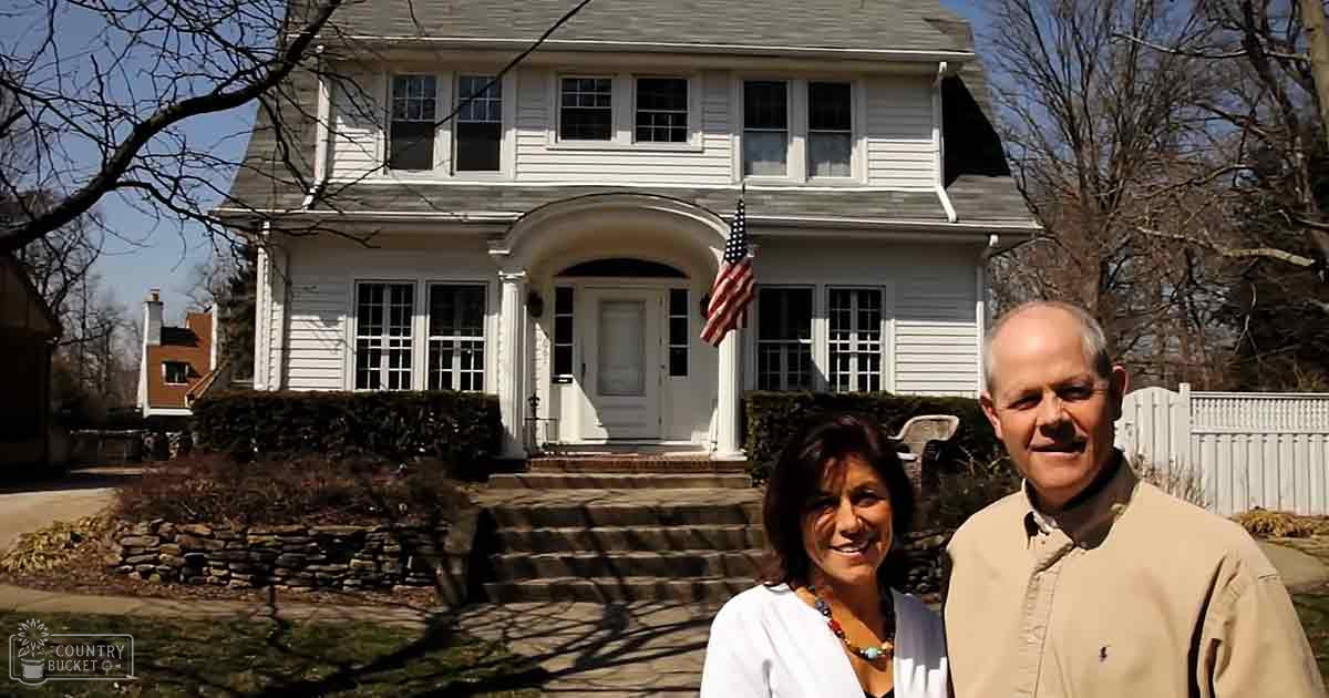 A couple stands casually in front of their suburban home, surrounded by a white picket fence and American flag, with bare trees suggesting springtime. The scene is illuminated by natural daylight.