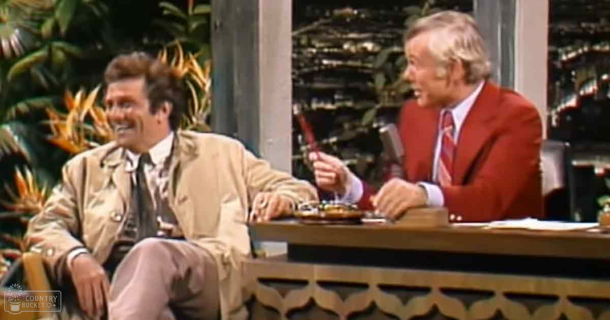 Adult male talk show host in beige trench coat, older adult male with red blazer, outdoor setting with wooden desk and plants.