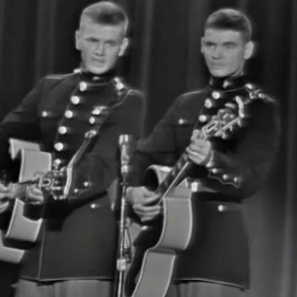 Two young adult male performers in mid-20th century military uniforms holding ceremonial swords, standing on an indoor stage with dark curtains and a microphone stand.