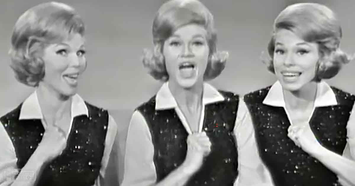 A group of performers on stage, dressed in mid-20th-century attire, possibly from a music video or theatrical production.