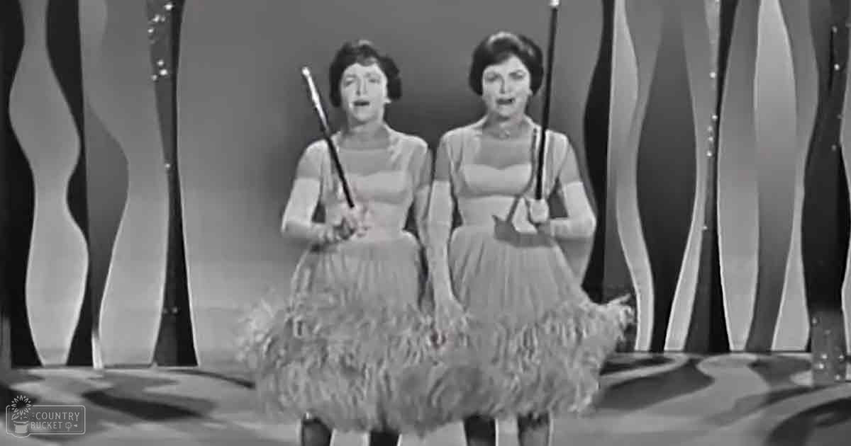 Two young adult female performers in stage attire with feathered skirts, likely from an older era, on a backdrop with wavy patterns and decorative elements.