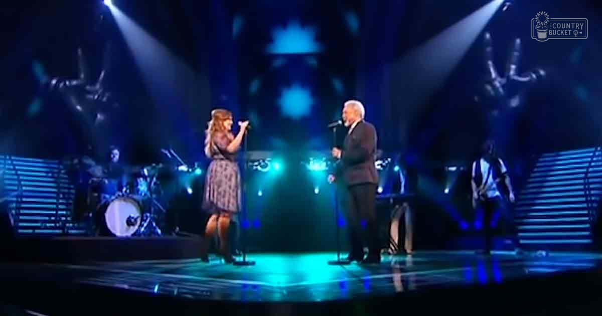 Indoor concert venue with a young adult female singer in patterned dress performing alongside an older adult male performer in formal attire, stage lighting and musical instruments visible.
