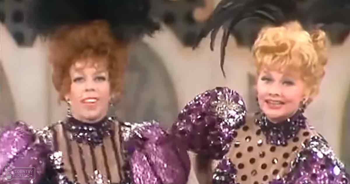 Two performers in vibrant costumes on a stage; adult female wearing purple sequined top with large hoop earrings, adult female in brown polka-dotted top with similar accessories, neutral indoor setting