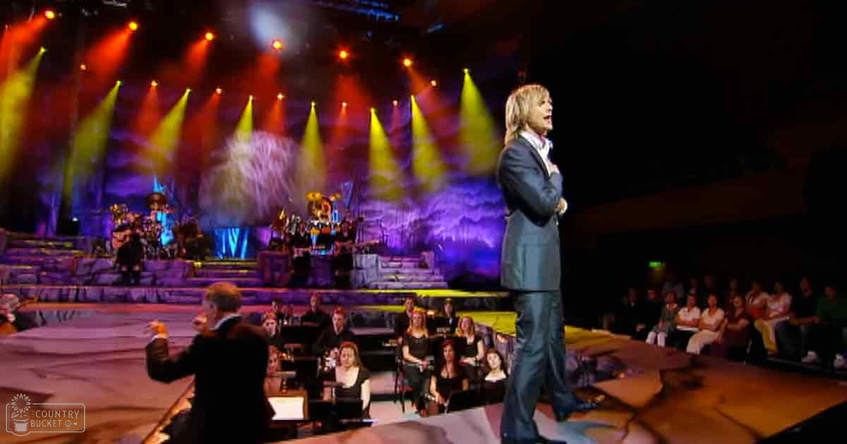 An adult male singer in a dark suit jacket on stage, accompanied by an older adult conductor and musicians playing stringed instruments under colorful spotlights in a dimly lit auditorium.