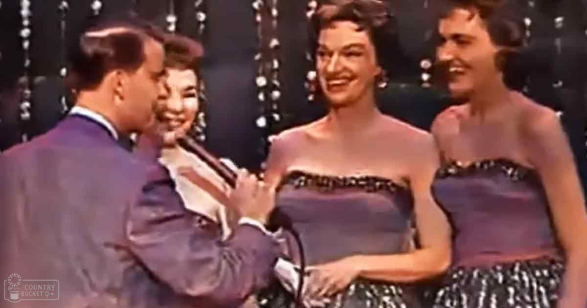 Three women wearing matching dark purple dresses with checkered hem details standing beside a man in a suit holding a microphone indoors at an evening event.