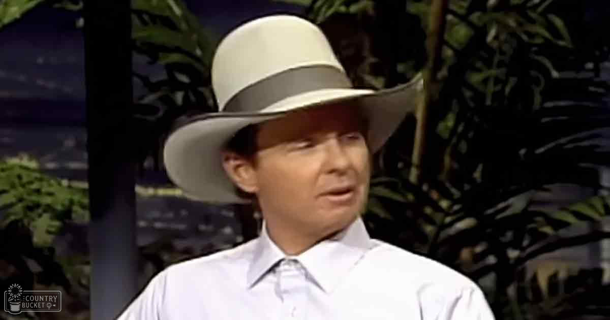 A young adult male with short dark hair wears a white cowboy hat with a gray band and a neatly buttoned white dress shirt in an indoor setting with artificial foliage in the background, suggesting it might be from a television show or studio.