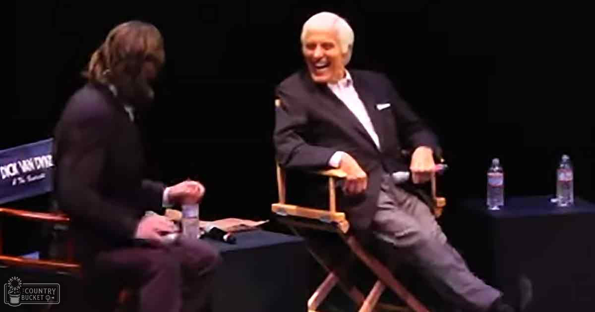 An older adult male singer with long hair, dressed in a dark suit and tie, seated on a director's chair at an event or talk show. Another older adult male is also seated nearby, wearing a light-colored shirt under a dark suit jacket.