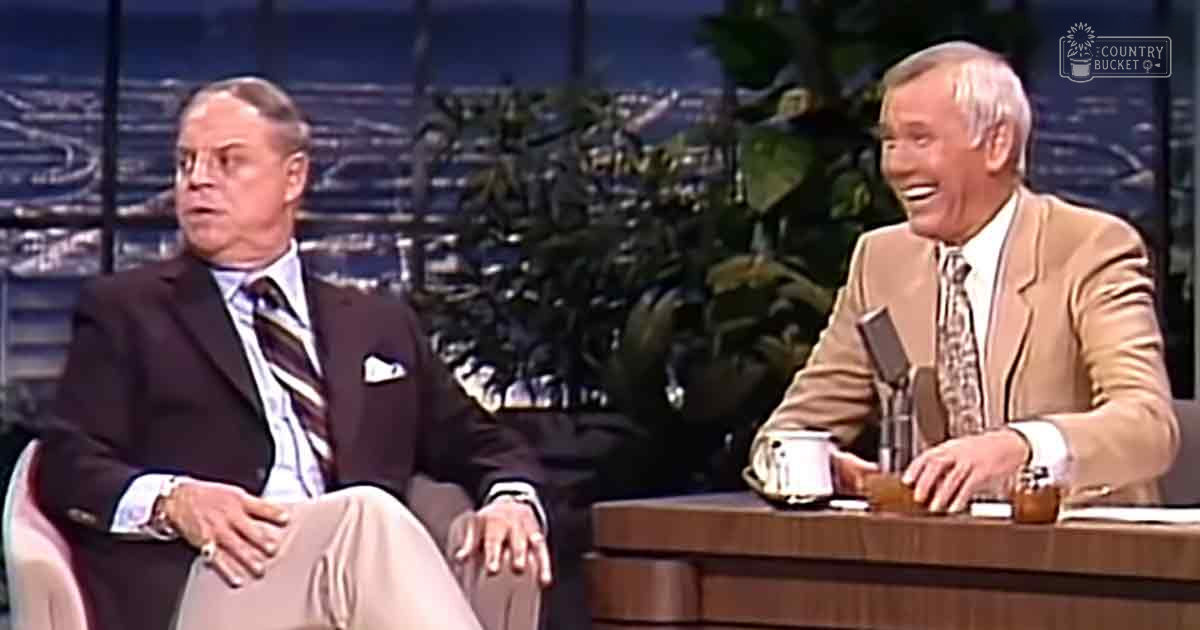 Two older adult men seated in a studio setting, one wearing a dark suit with a striped tie and the other in a tan blazer over a patterned tie, discussing against a backdrop of greenery and cityscape visible through large windows.