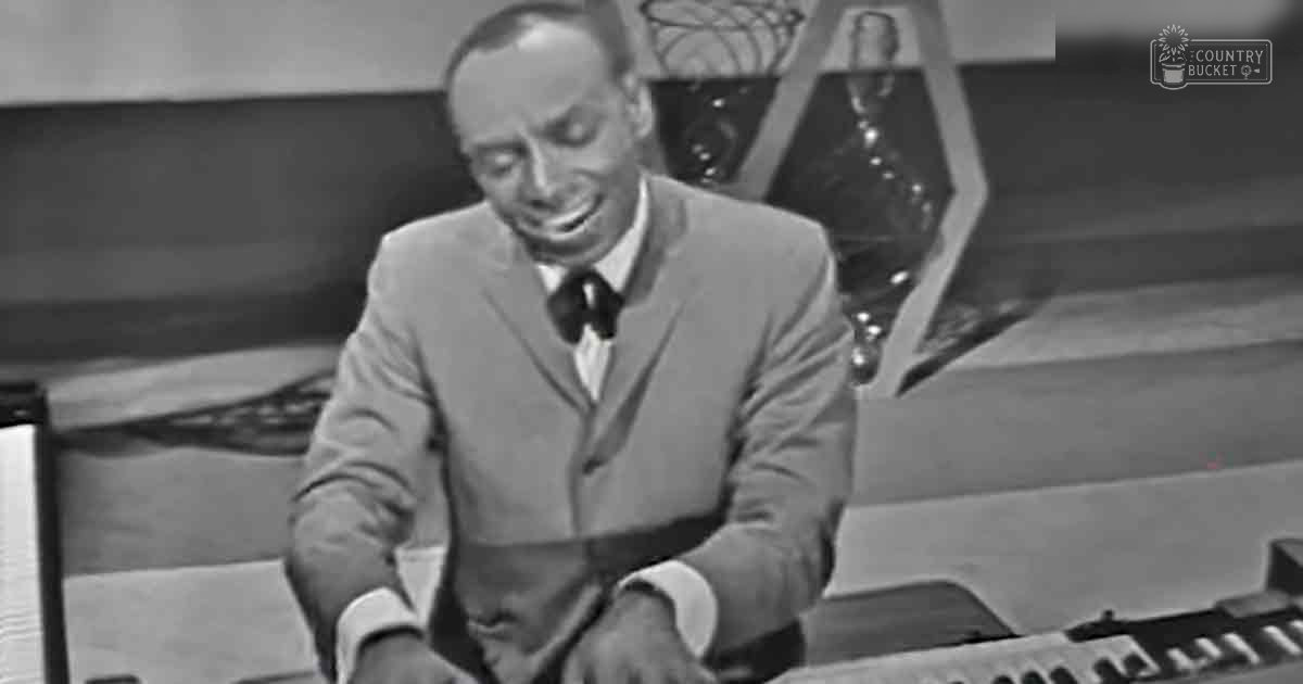 An adult male wearing a tailored suit jacket with a tie, playing an organ in a mid-20th century studio setting.