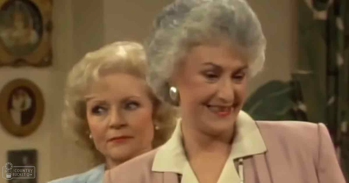 An older adult woman in a light pink blazer with vertical stripes, standing indoors near another older adult female in a light blue patterned jacket.