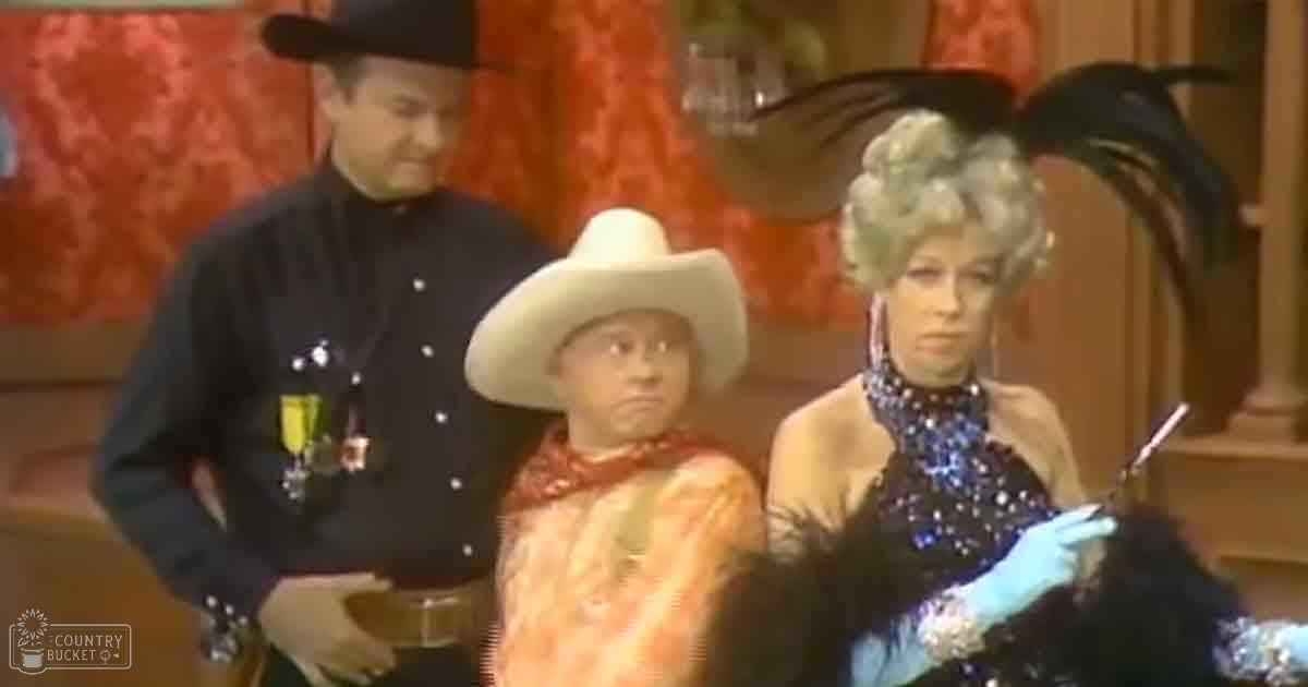 A comedic sketch scene with characters dressed in Western attire, set indoors against red curtains, under bright lighting.