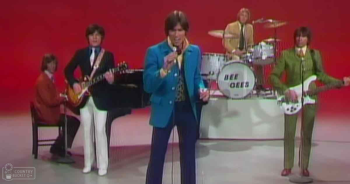 A male adult singer in a bright blue striped suit and yellow shirt stands center stage with other musicians. A young adult male guitarist plays an electric guitar to the left, while another adult male drummer sits on the right playing drums. A fourth adult male performer holds a guitar at the far right. The backdrop is solid red with soft lighting, suggesting an indoor music studio setting.