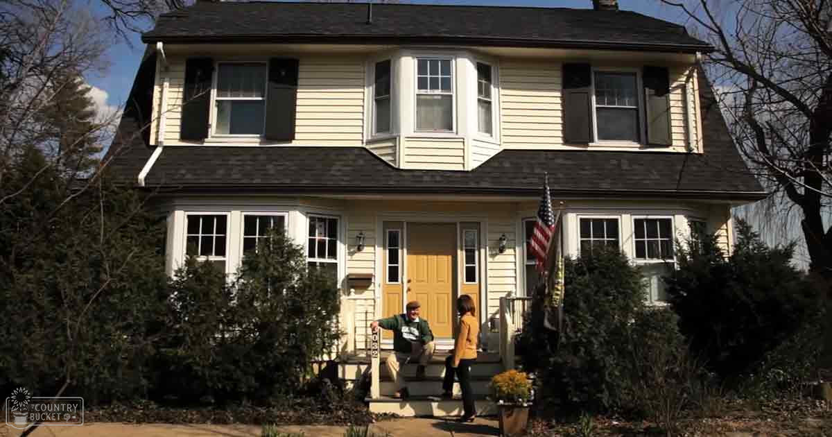 Two individuals stand in front of a house with yellow doors and white trim, surrounded by bare trees suggesting late fall or early spring. The scene is set under clear blue skies during the daytime.