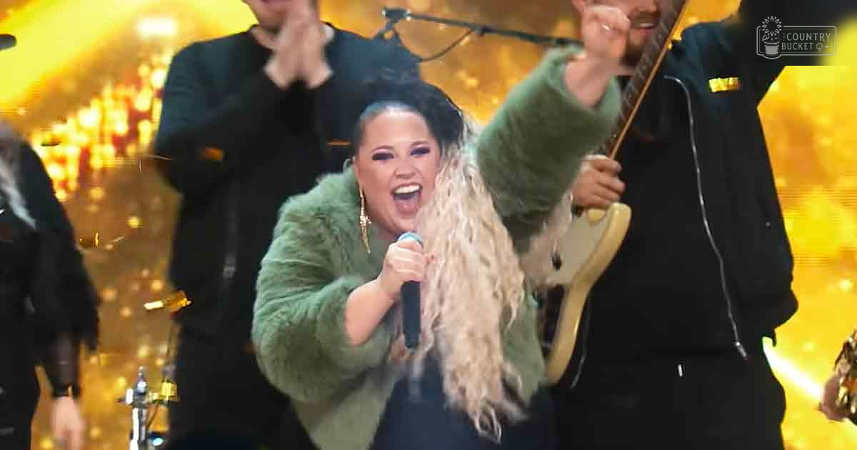 Adult female singer wearing a green fuzzy jacket performs on an indoor concert stage with bright golden lighting, accompanied by male musicians holding electric guitars.