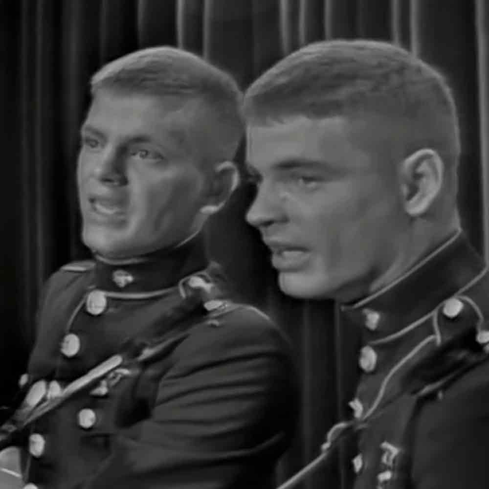 Two young adult males in military uniforms stand against a dark curtain backdrop, suggesting an indoor setting for a performance or ceremony.