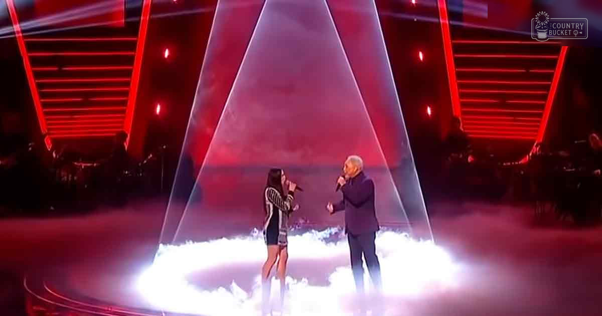 A young adult female in a black and white patterned dress performs on stage with a middle-aged male in a dark suit, set against red lighting and geometric light beams, suggesting a performance or competition at night.