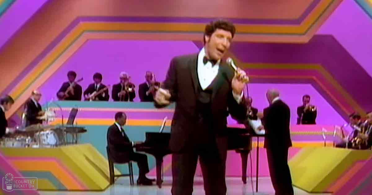 An adult male singer in a black tuxedo with a white shirt and bow tie performs on stage, accompanied by an orchestra of middle-aged to older adults in dark suits.