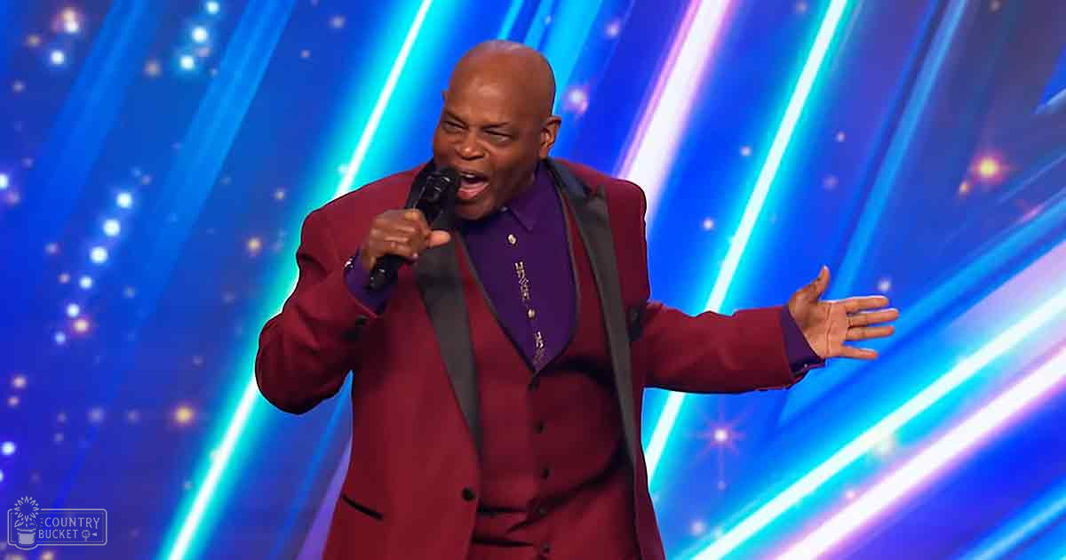 An adult male performer in a modern formal maroon suit jacket with long sleeves on stage under dynamic blue and white lighting.