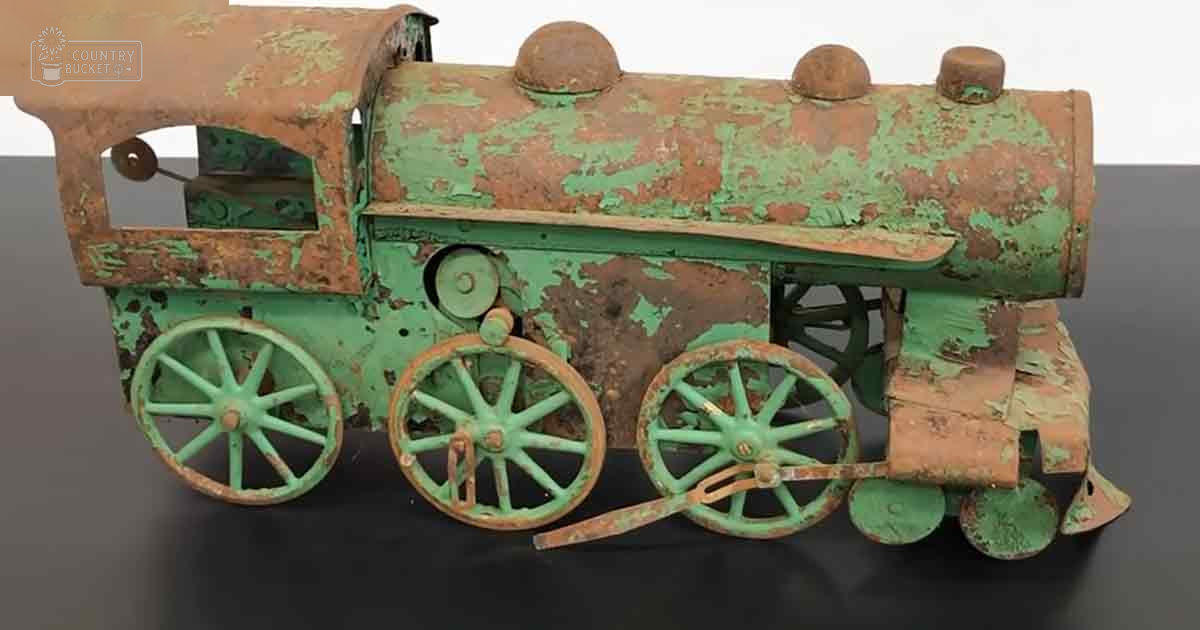 An old toy steam locomotive with green and rust-colored paint sits indoors, showing signs of age on a dark surface.