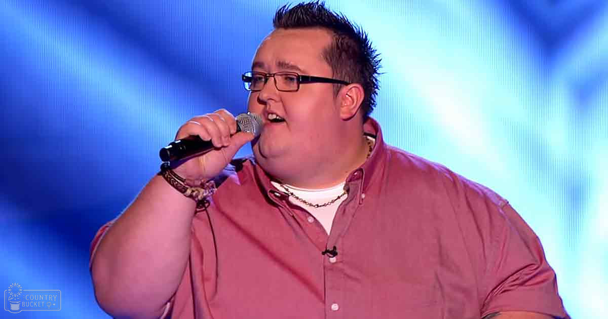 Young adult male in a relaxed, casual pink button-up shirt with glasses and bracelets on stage under blue lighting.