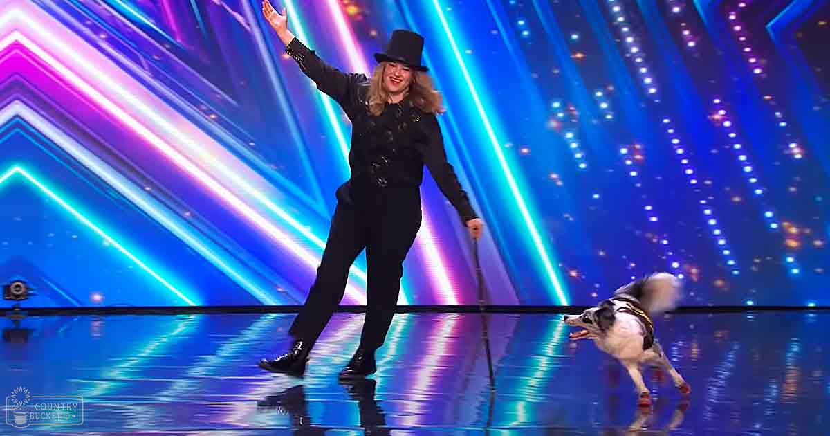 An adult female performer in a talent show with her Border Collie, dressed elegantly and accessorized with a tall black hat, set against a vibrant stage backdrop.