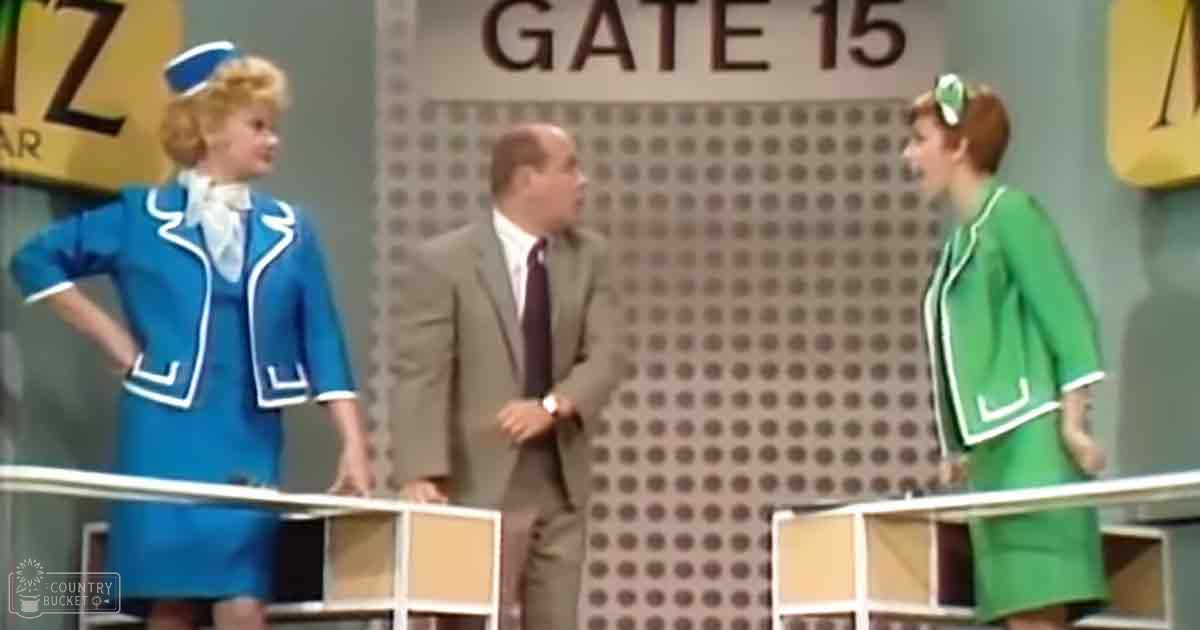 A man in his 40s or 50s wearing a light brown suit jacket over a white shirt and dark tie, standing on stage with two flight attendants in blue and green uniforms. The setting is indoors, likely for a performance or talk show.