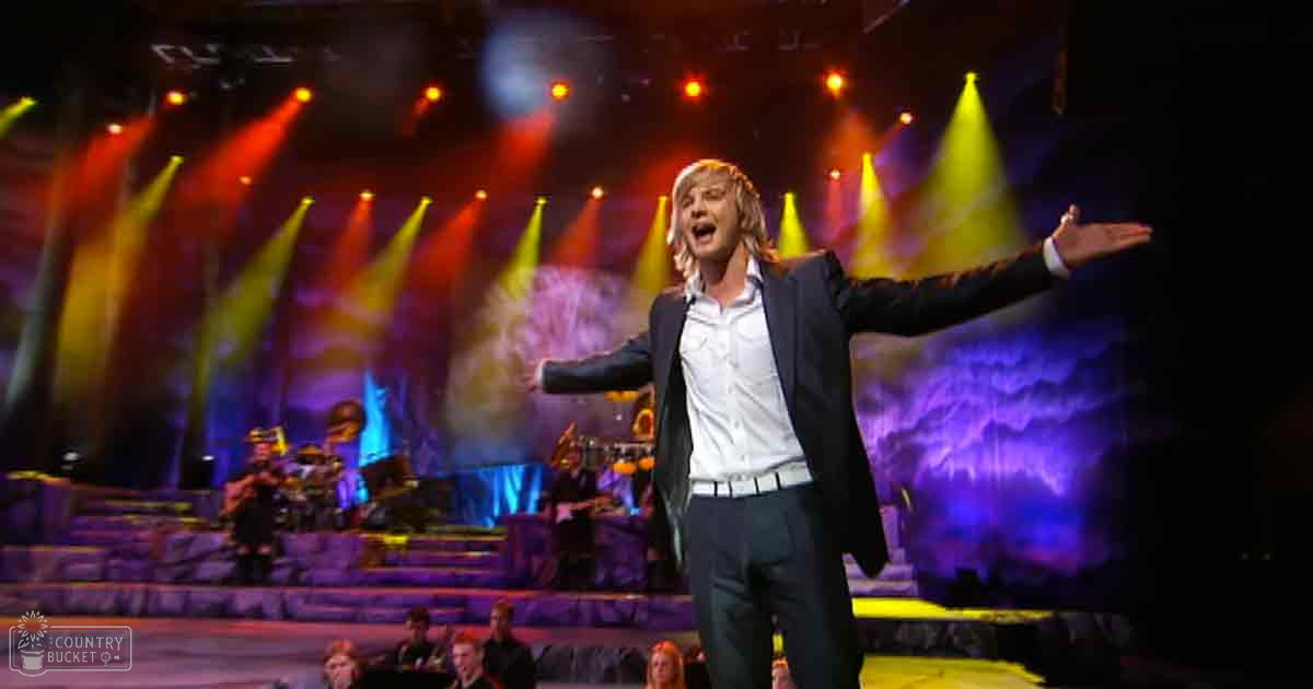 A middle-aged male performer in a concert setting with colorful stage lighting and an audience seated on tiered seating within an indoor venue.