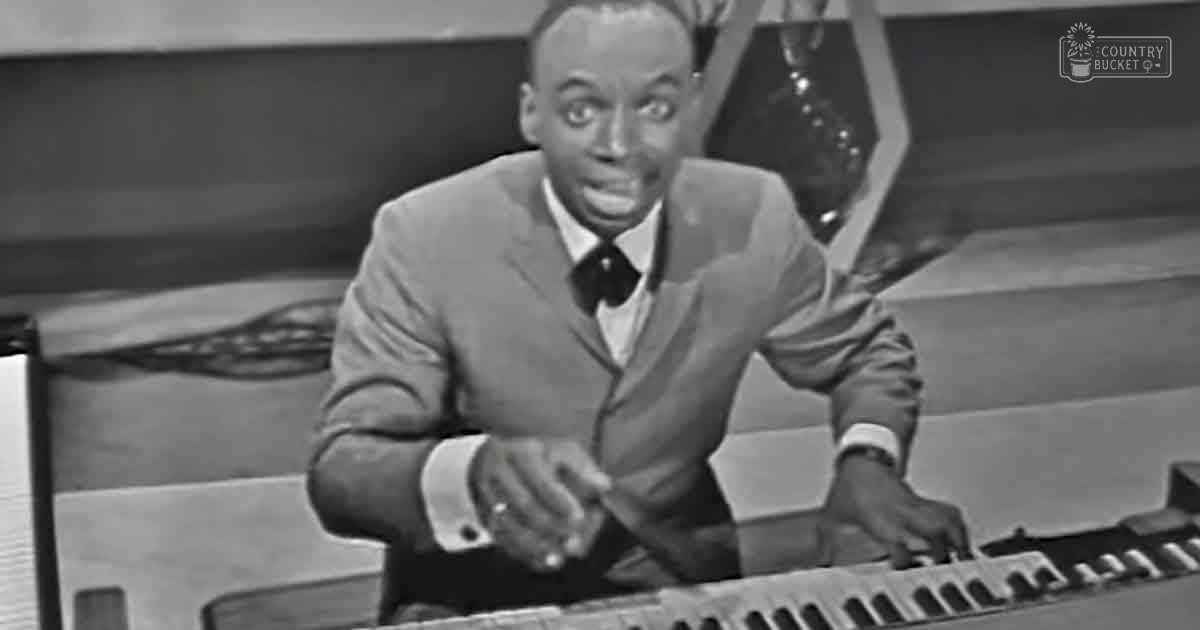 Man in adult age band performing at music studio or stage setting with light-colored suit jacket, white shirt, dark bow tie and keyboard or piano in foreground; ornate trophy-like object on stand in background;