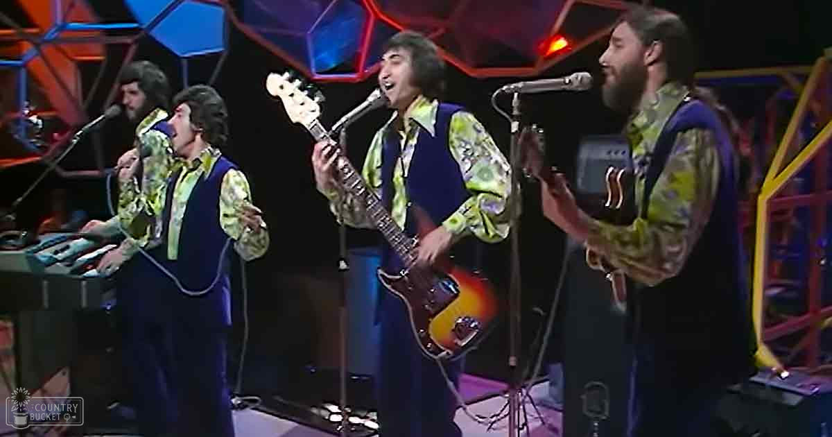 An indoor musical performance with adult males in floral-patterned shirts and electric guitars, set against a vibrant geometric backdrop.