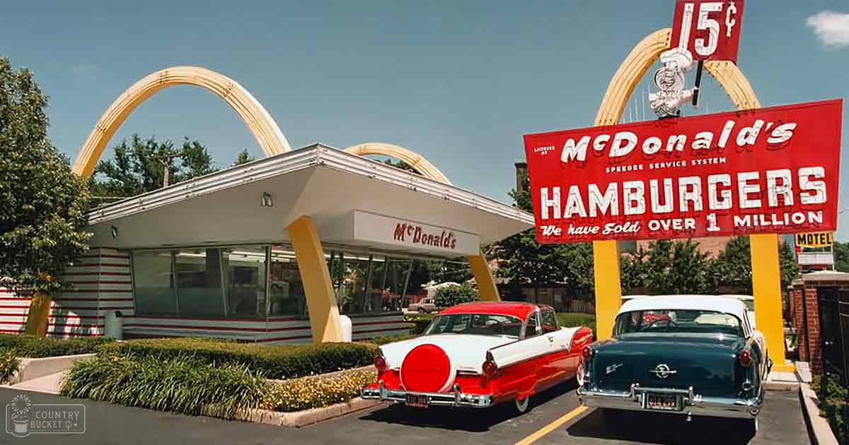A vintage McDonald's restaurant under clear skies with two classic cars parked in front, iconic golden arches above, and lush greenery surrounding it.