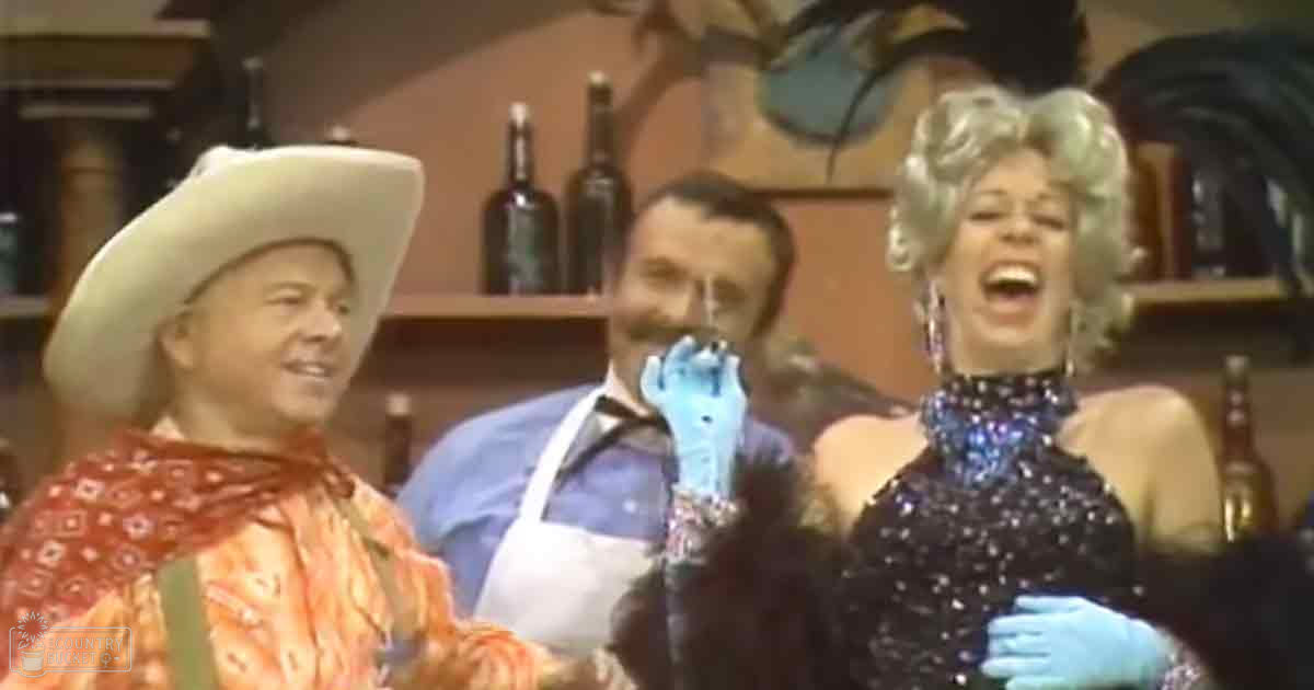 An older adult woman in a black sequined dress with large earrings stands beside an adult male wearing a red bandana and cowboy hat, while another middle-aged man in blue gloves and apron is present. The setting appears to be a bar or restaurant with shelves of bottles and decorative items.