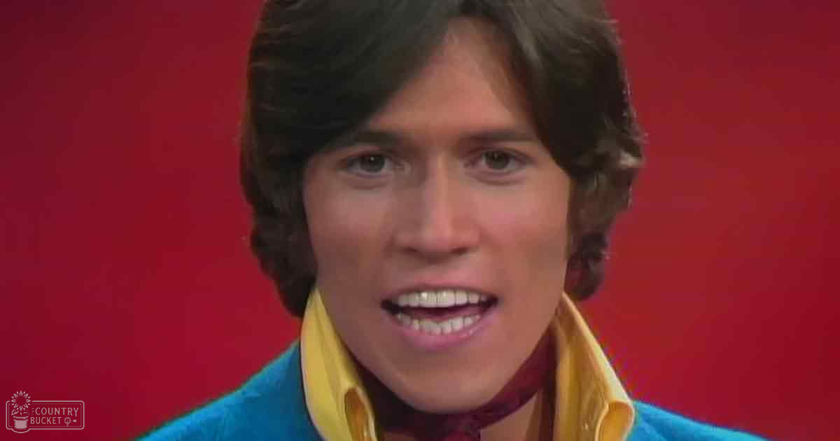 Young adult male wearing a high-collared, relaxed fit collared shirt with blue color and yellow accents indoors against a red background.