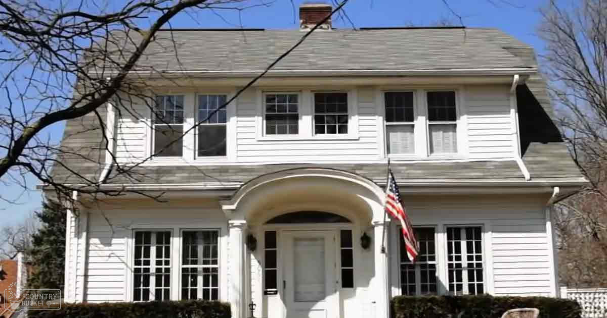 A white two-story house with a gray roof, an American flag on its front porch, located in a suburban neighborhood. Bare tree branches suggest spring or summer.