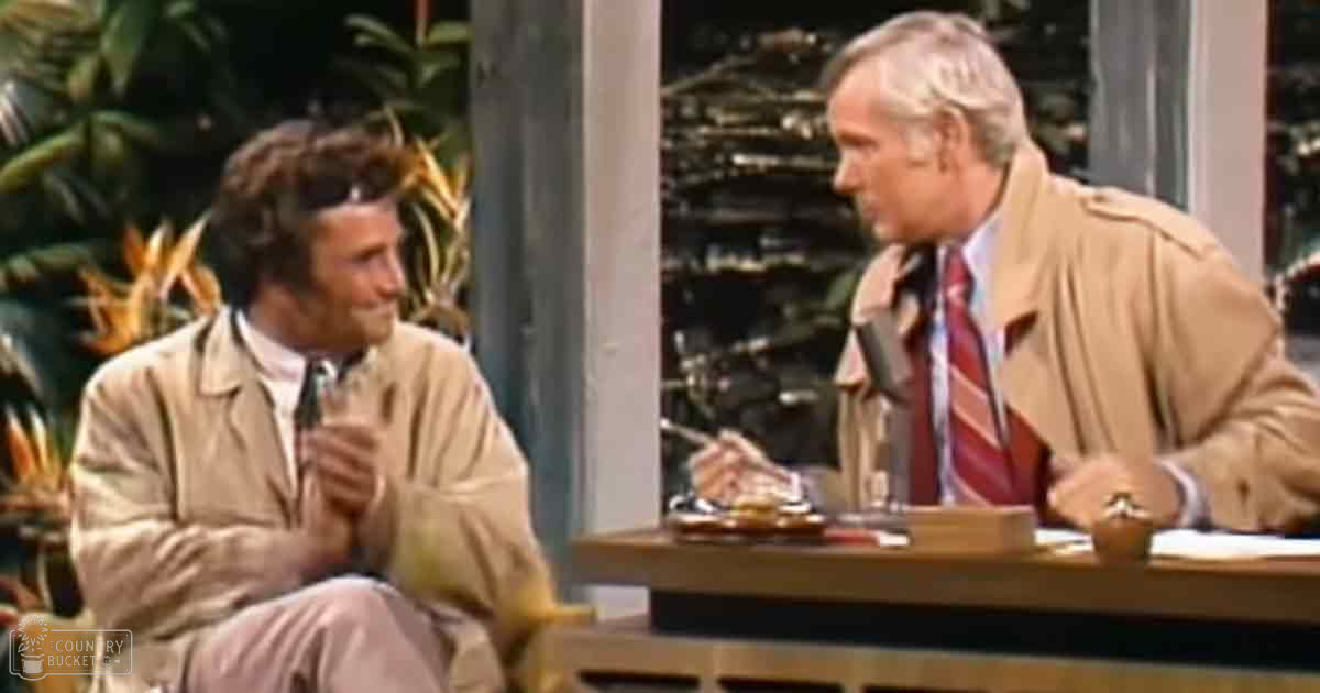 A young adult male and an older adult male, both wearing beige trench coats over formal shirts with patterned ties, seated at a wooden desk with microphones in a professional studio environment.
