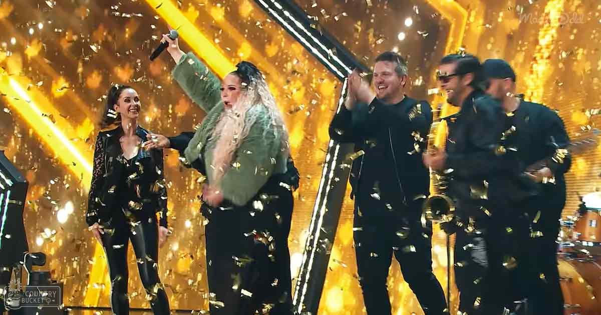 Adult female in a black jumpsuit with gold accents at an awards ceremony, alongside other performers and microphones on stage.
