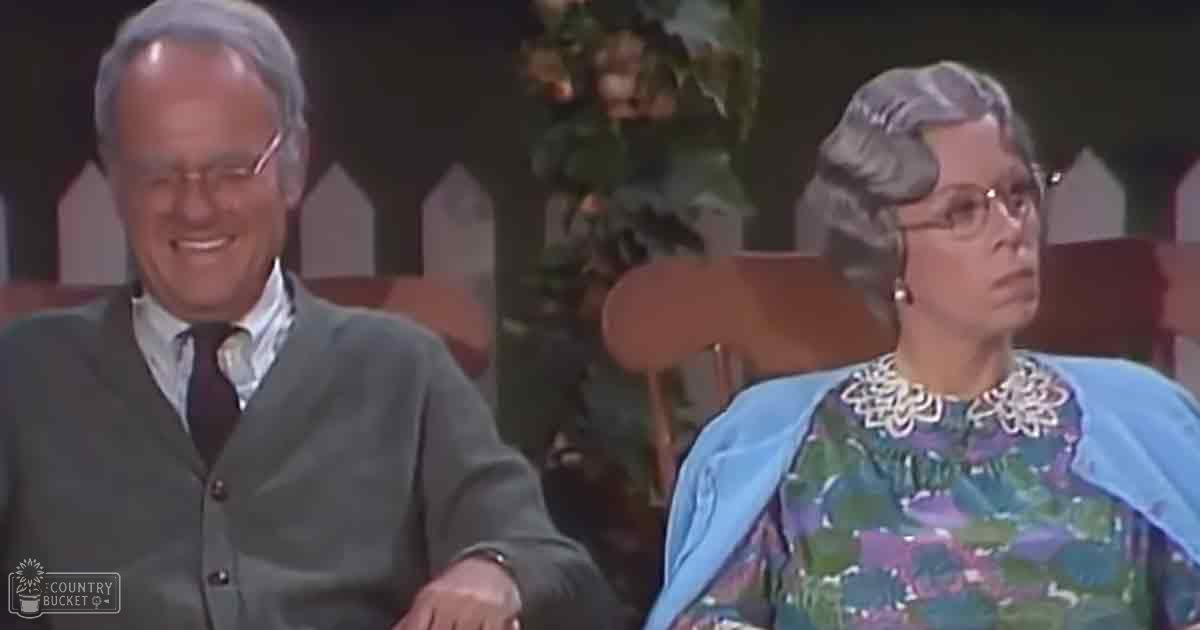 An older adult male and female seated together, wearing formal attire typical of the mid-20th century in a television studio setting with decorative elements.