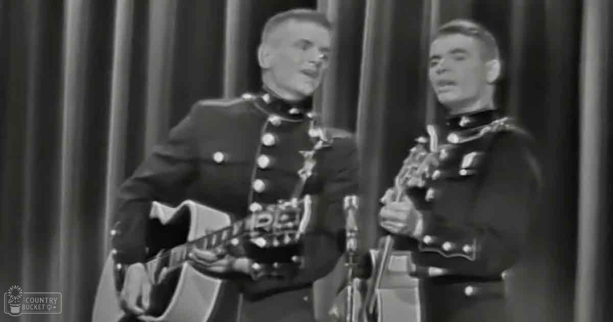 Two adult male singers in historical military uniforms with rifles, performing indoors against a backdrop of curtains.