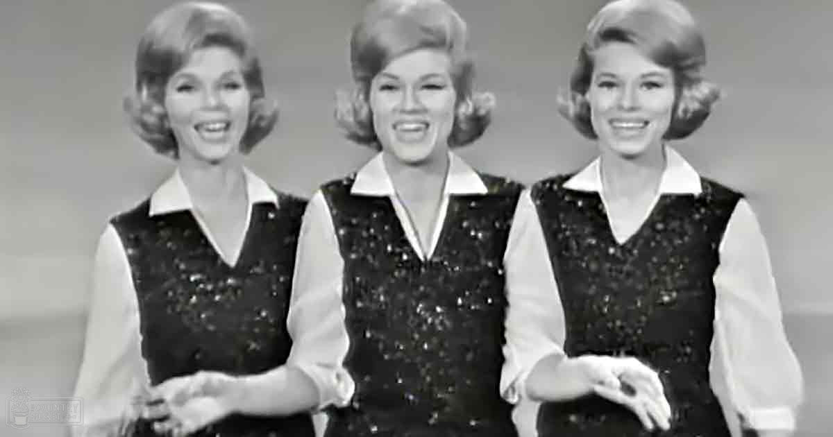 Three women performing in a studio setting, dressed identically in white blouses with black vests adorned with small decorative elements or sequins.