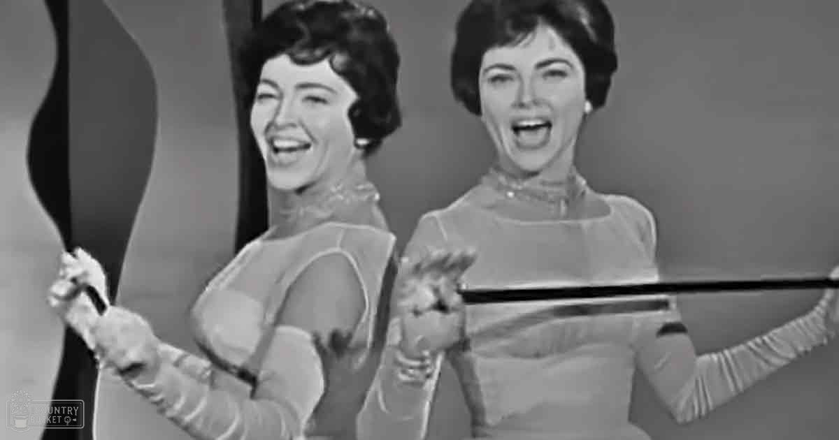 Two young adult females wearing shiny, metallic-patterned tops with gathered wrists, holding violin bows in a studio setting with neutral backdrop.