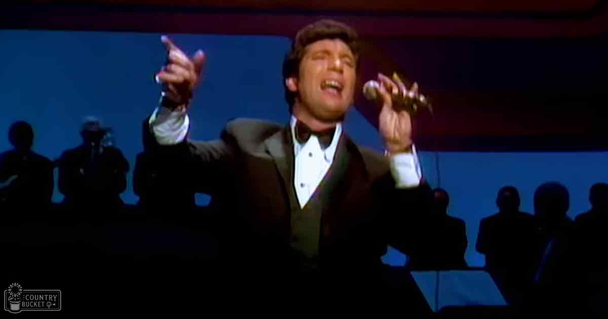 Adult male performer in a dark suit with white shirt and black bow tie, holding a microphone on an indoor stage setup at night.