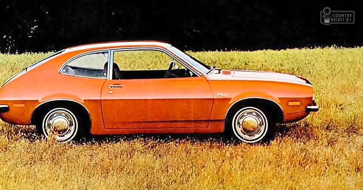An orange vintage sports car parked in a field, tall grass suggests spring or summer daylight natural lighting