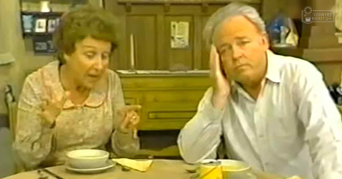Two older adults dining in a restaurant setting with wooden decor and framed pictures on the wall, wearing light-colored garments with floral patterns for one and button-down shirts for the other.