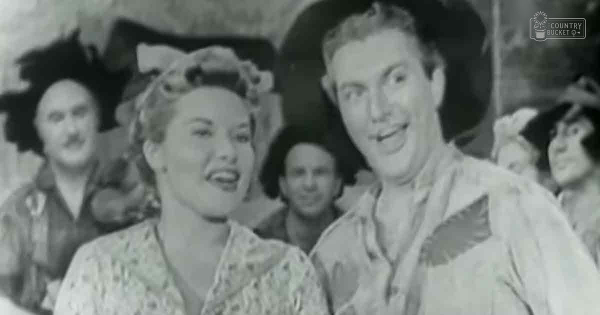 A social gathering with a couple smiling indoors under natural daylight, surrounded by blurred figures and a mix of floral-patterned garments.